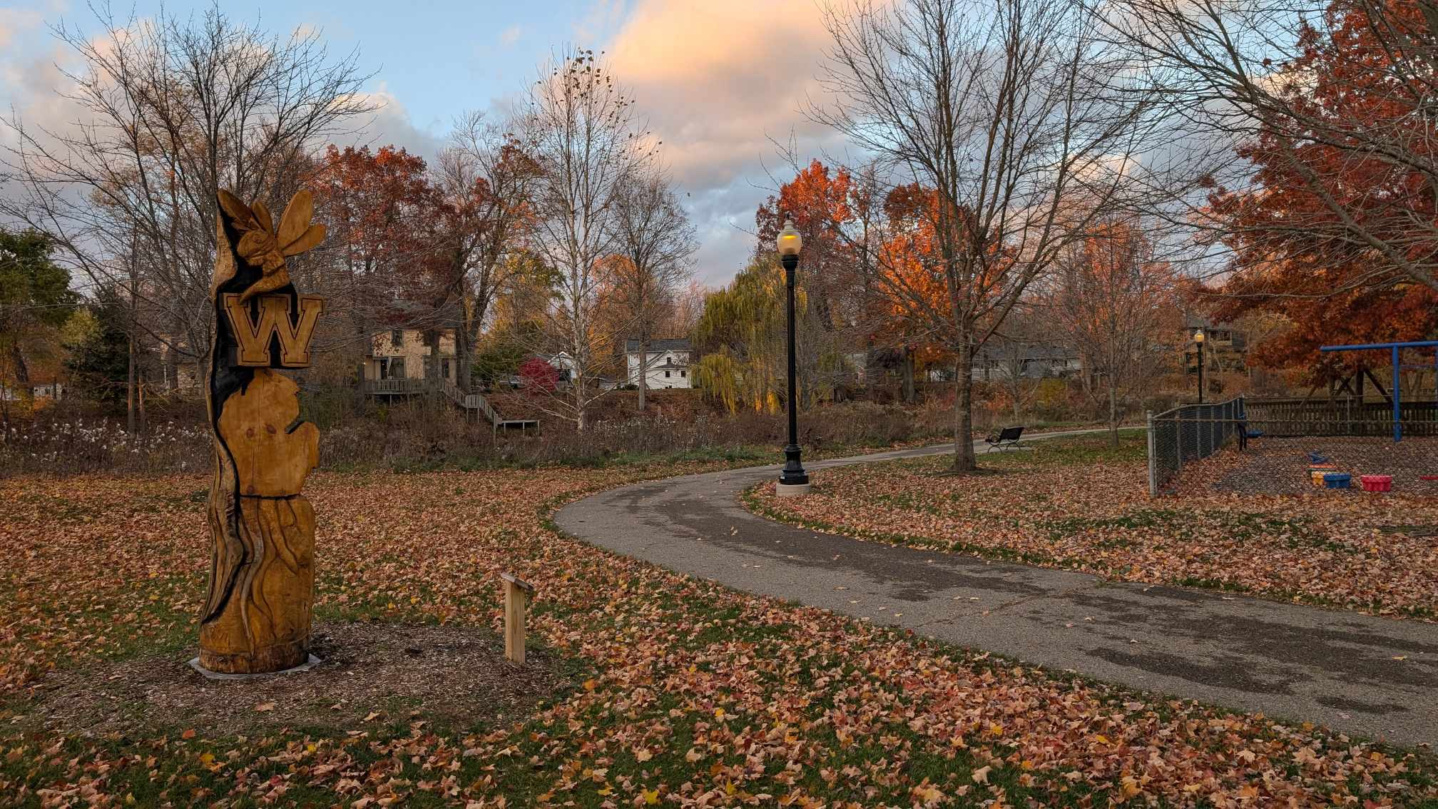 Walking path at McCormick Park in downtown Williamston, Michigan, surrounded by autumn trees and a Willy tree sculpture near the Putnam bridge.