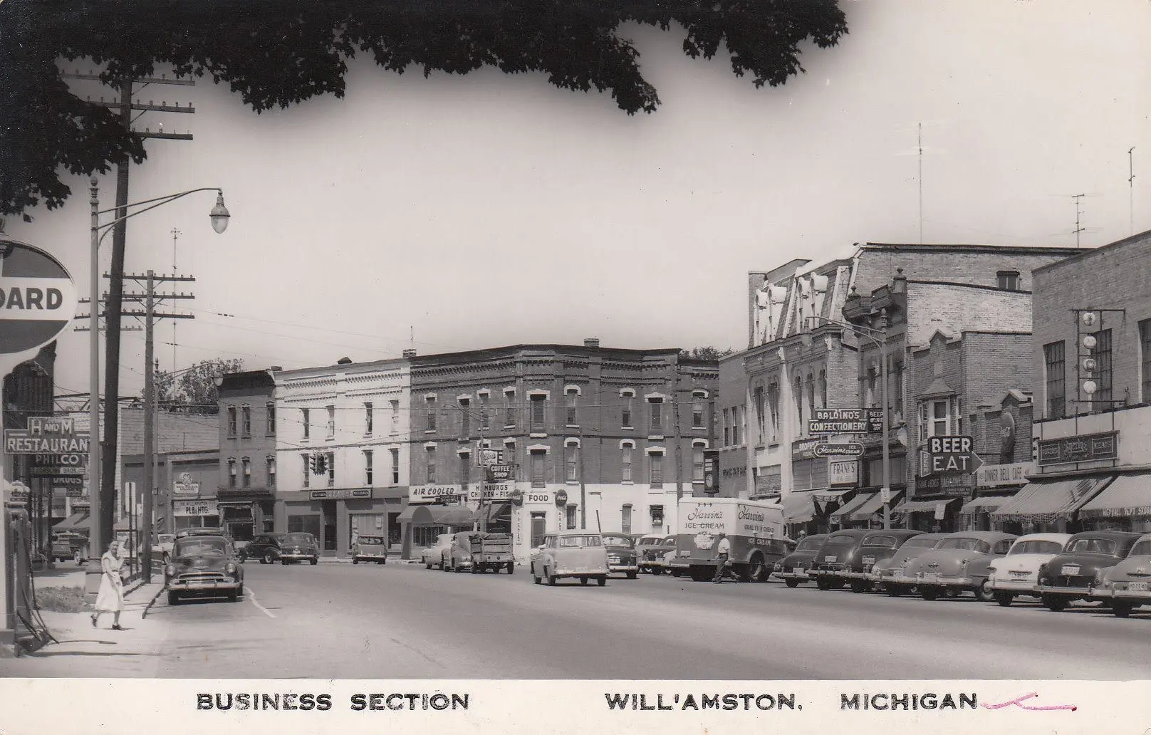 Historic downtown Williamston, Michigan, showing 1950s-era storefronts, vintage cars, and small-town businesses along Grand River Avenue.